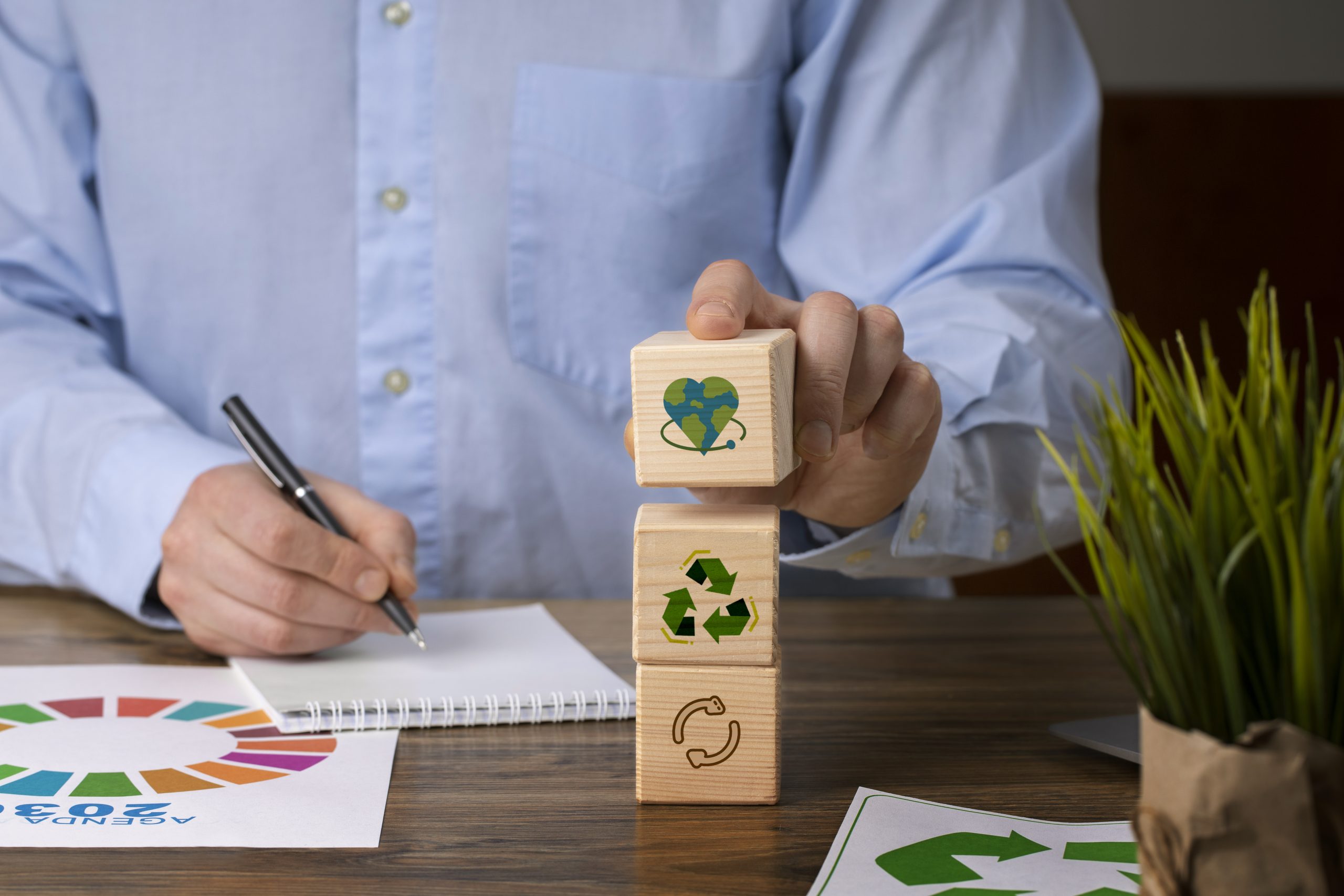 A person in a blue shirt stacking wooden blocks with sustainability icons—recycling, circular economy, and Earth—on a desk with ESG-related documents and a green plant, symbolising ESG investment planning and sustainable business strategies.